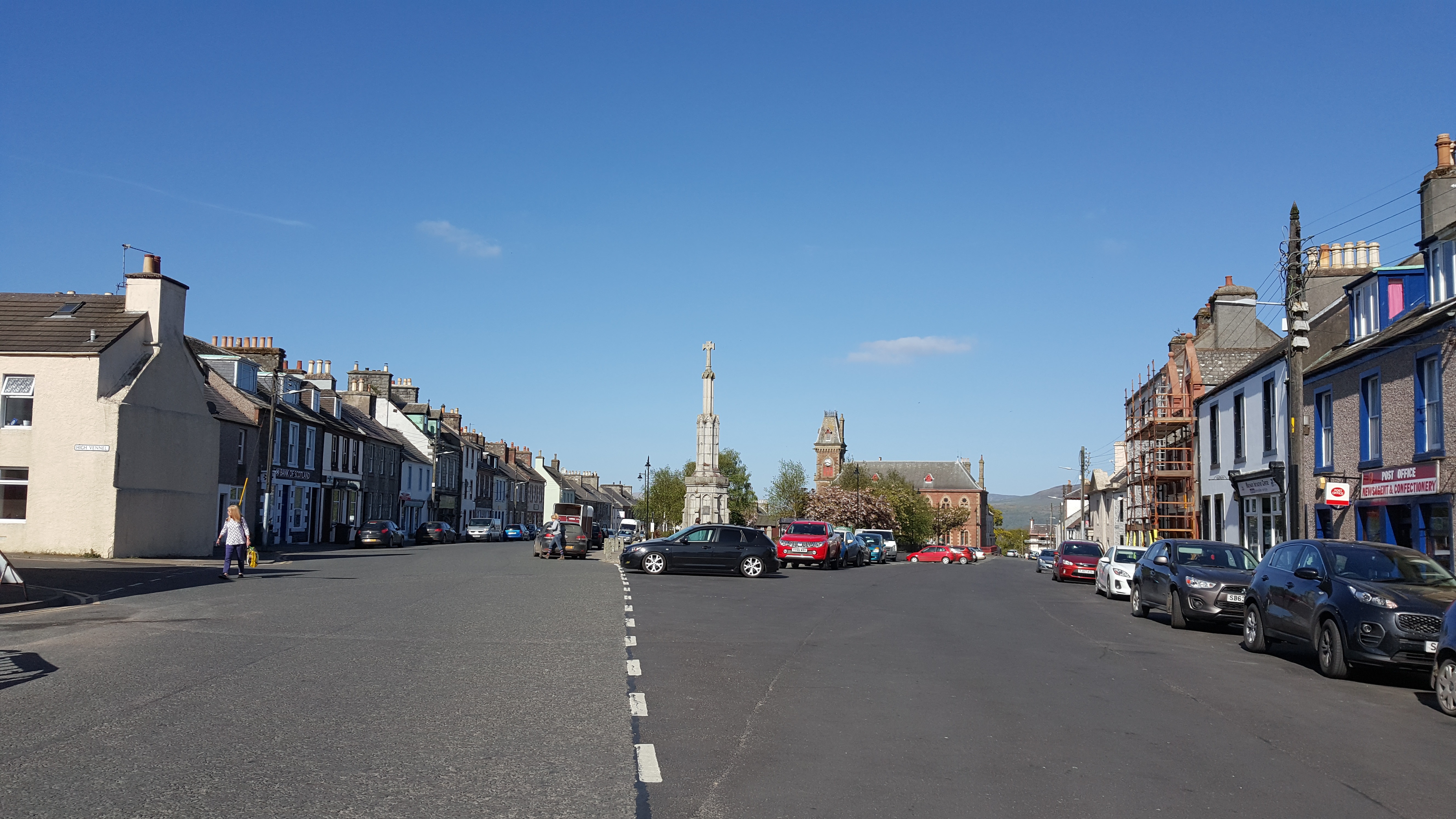 Wigtown main street lined with cars, terraced houses and shops. Mercat Cross and the County Buildings in the distance.
