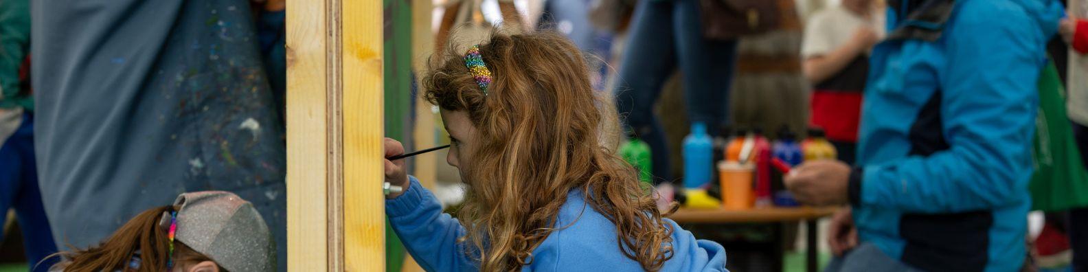 At a children's art workshop, a young child uses a paintbrush on a large board. In the background are bottles of paint and brushes.