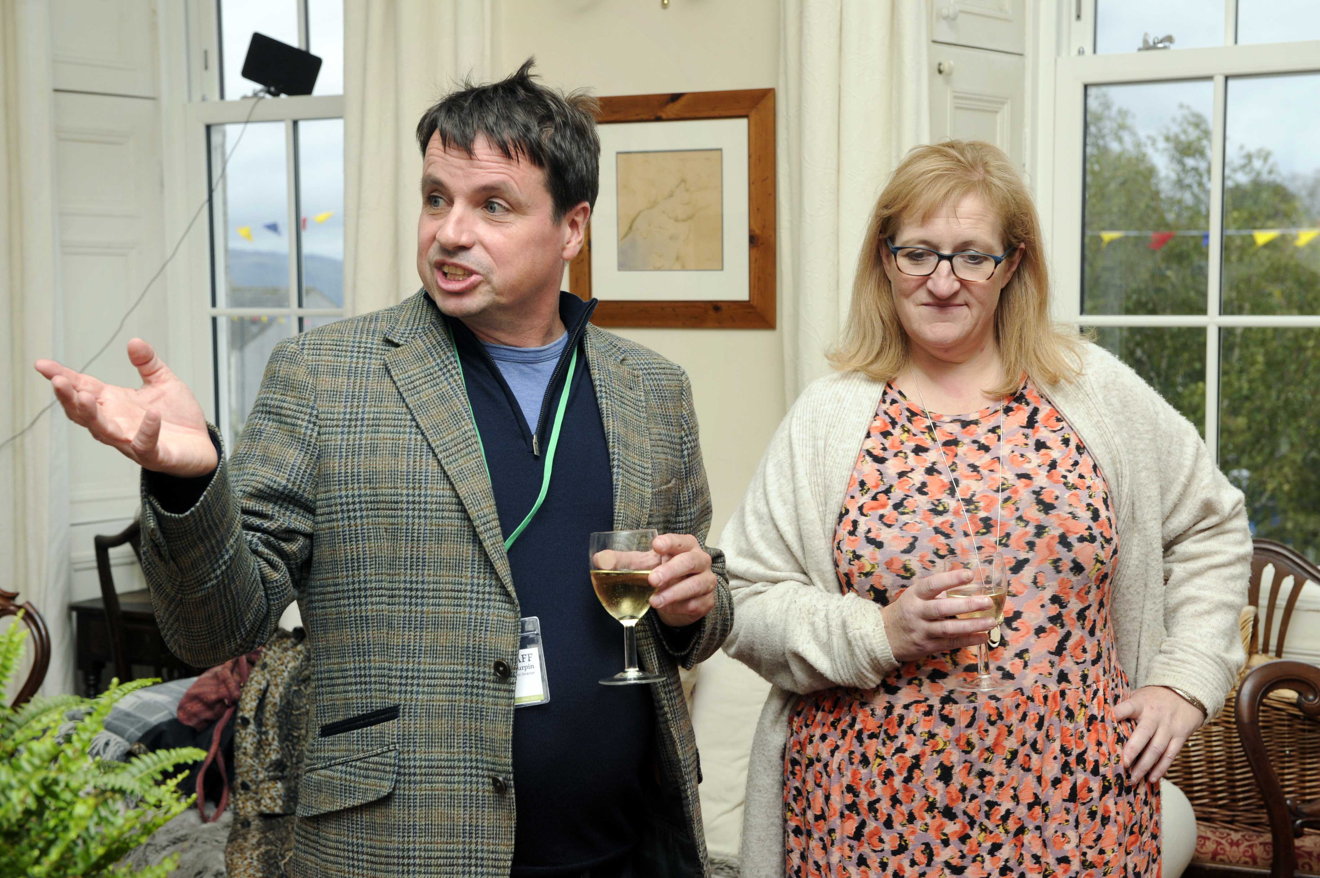 Adrian Turpin and Jo Lawrence standing in Craigard Gallery during the Wigtown Book Festival. They each hold a glass of wine, Adrian is gesticulating with his hand.