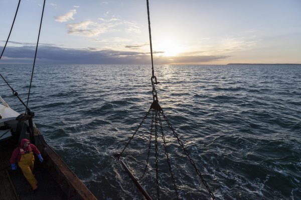 The lowering of the scallop dredger on a fishing vessel. The sun is setting in the distance.
