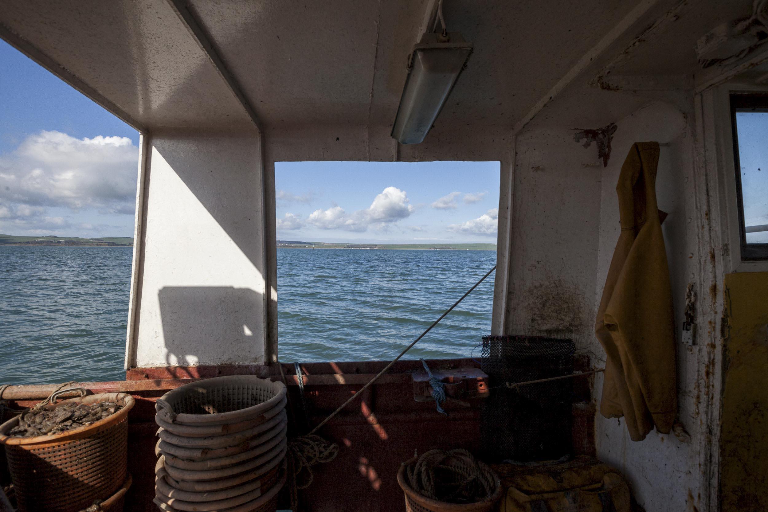 Inside the hull of a fishing vessel. Empty buckets are stacked up, one contains fish. A yellow waterproof coat is hanging up.