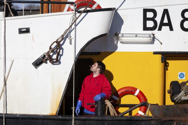 A person is standing on the deck of a fishing vessel smoking in the sunshine.
