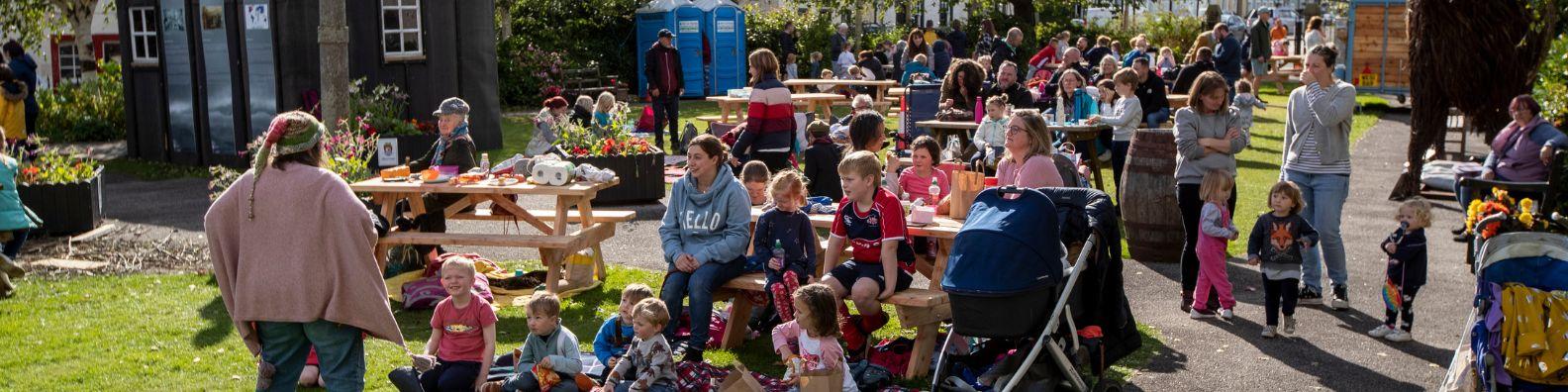 At an outdoor children's event during Wigtown Book Festival. A group of young children sit in the sunshine on grass and at picnic tables as they are entertained by Renita Boyle.