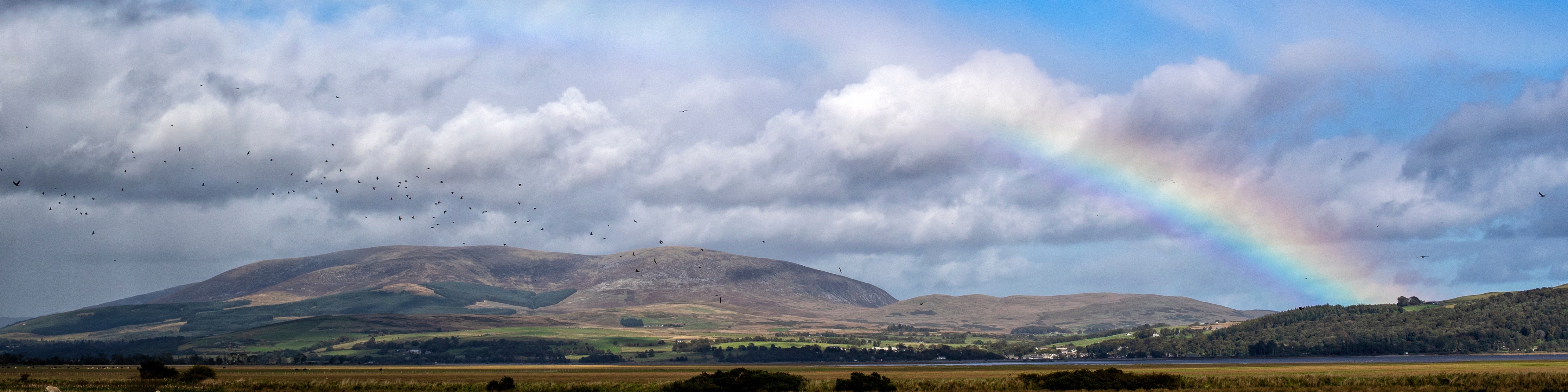 View of the Cree Estuary and Galloway hills. The blue sky is full of low rain clouds, a rainbow is present.