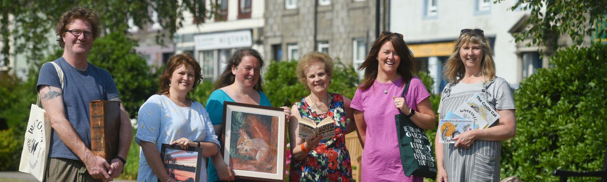 During the Wigtown Book Festival Cathy Agnew, Shaun Bythell and stall holders from The Kist stand together in Wigtown gardens. They are holding books and pictures.