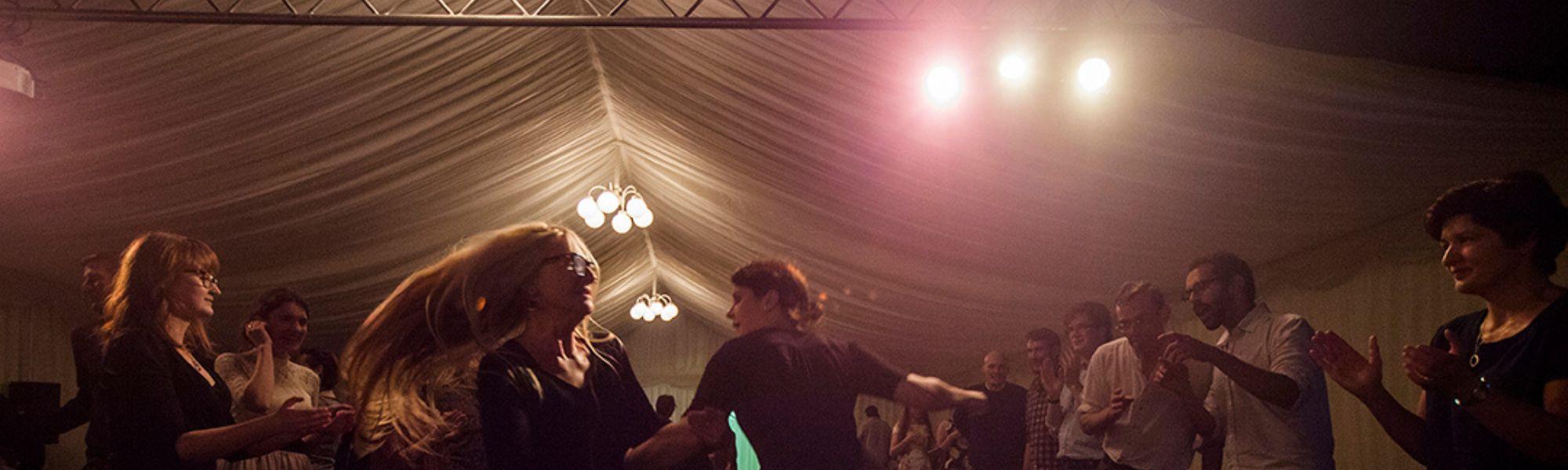 A group of people dancing and clapping at the Wigtown Book Festival Ceilidh. Bright lights are shining above them.