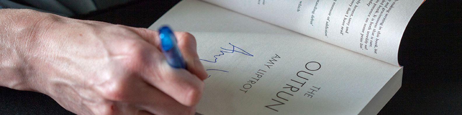 A hand holding a blue pen writing on the inside cover of an open book.