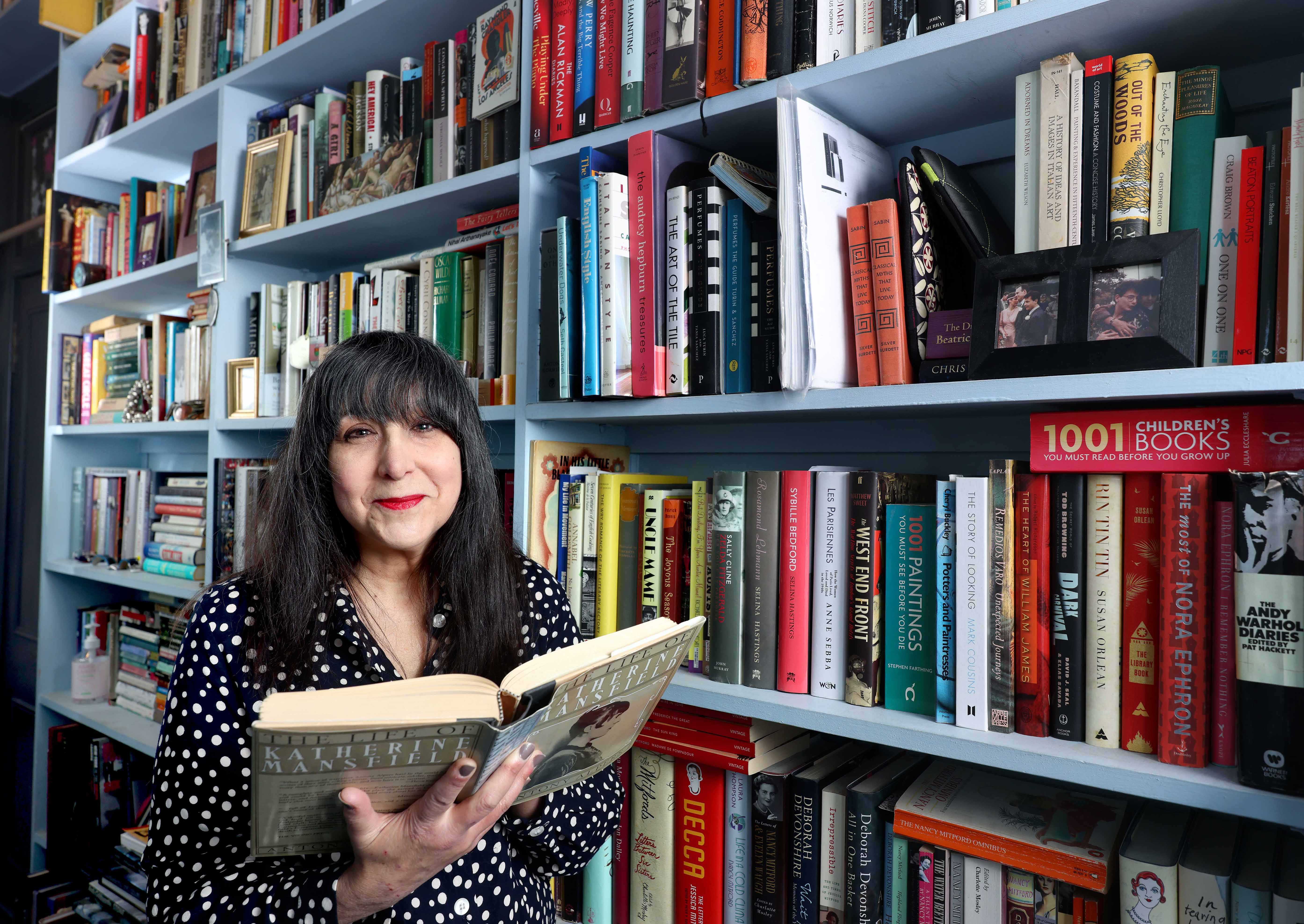 Lee Randall stands in a bookshop smiling and holding open a book. The bookshelves behind her are full of books.