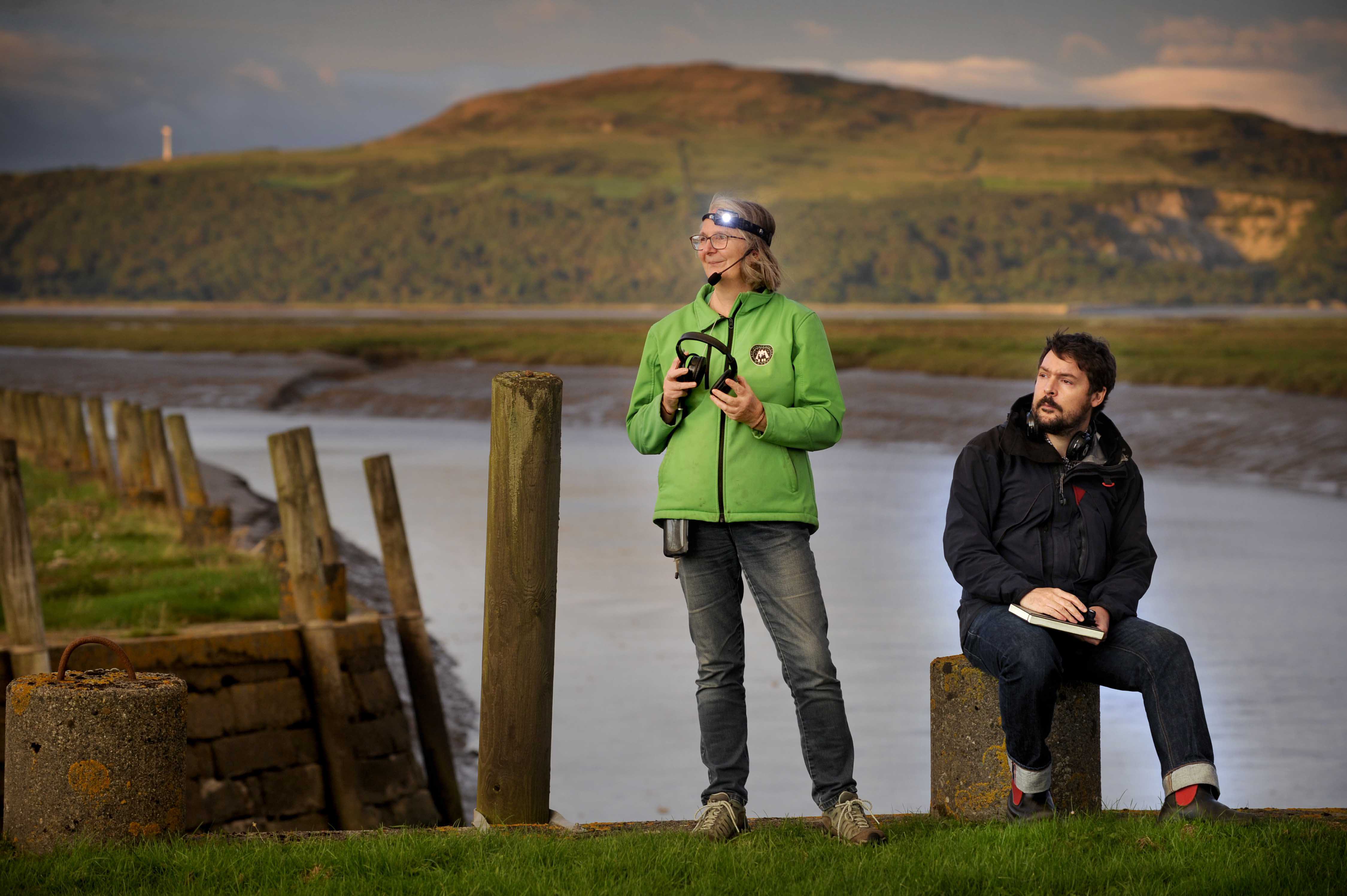 Elizabeth Tindall and Dan Richards standing at Wigtown Harbor during their event at Wigtown Book Festival.  They are both wearing headphones, Elizabeth is also wearing a head torch and microphone. The sun is setting on the hills behind them.