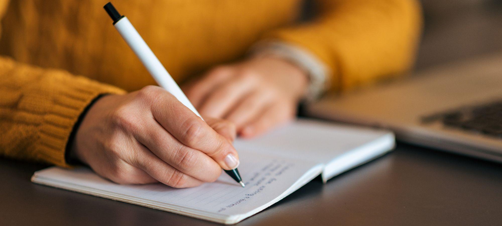 A person is sitting at a desk with a pen in their right hand writing in an open notebook.