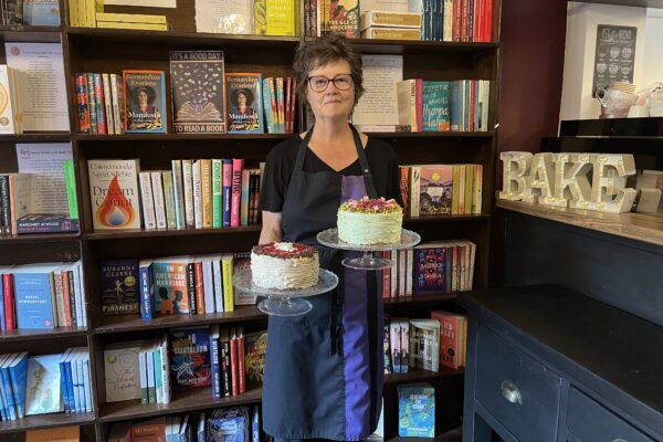 Jacqui owner of ReadingLasses holding two round cakes in front of a vibrant bookcase