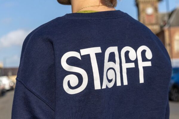 Image of Wigtown Book Festival navy blue sweatshirt, reading 'Staff' across the back. The staff member wearing the jumper faces away from the camera, pink dyed hair tied in a bun is visible at the top of the frame. Wigtown County Buildings is blurred in the background.