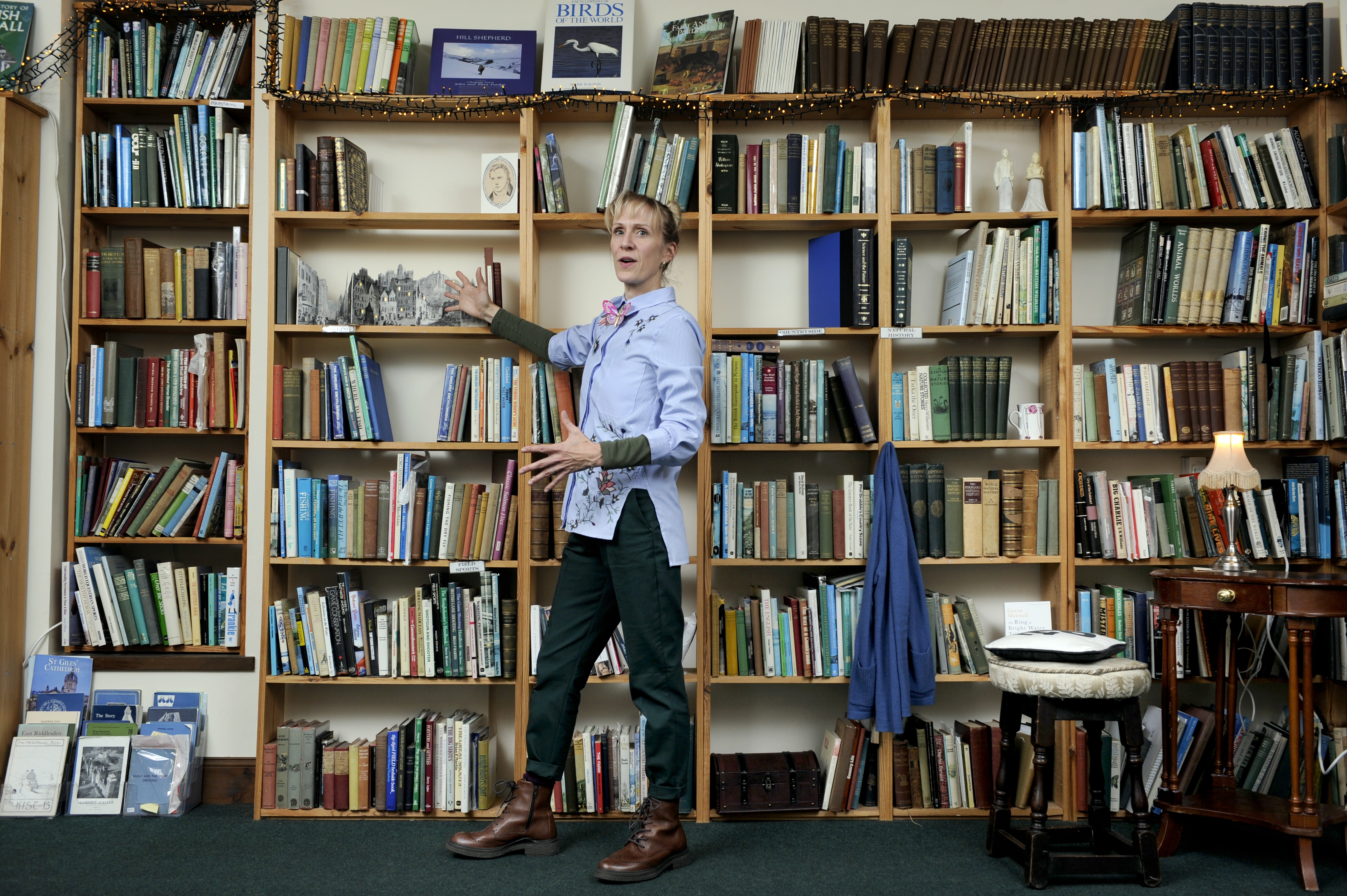 A person is standing in a bookshop. They are pointing towards a picture surrounded by bookshelves full of colourful books. Fairy lights adorn the top shelf, a small table holds a lamp and a stool sit in the corner.