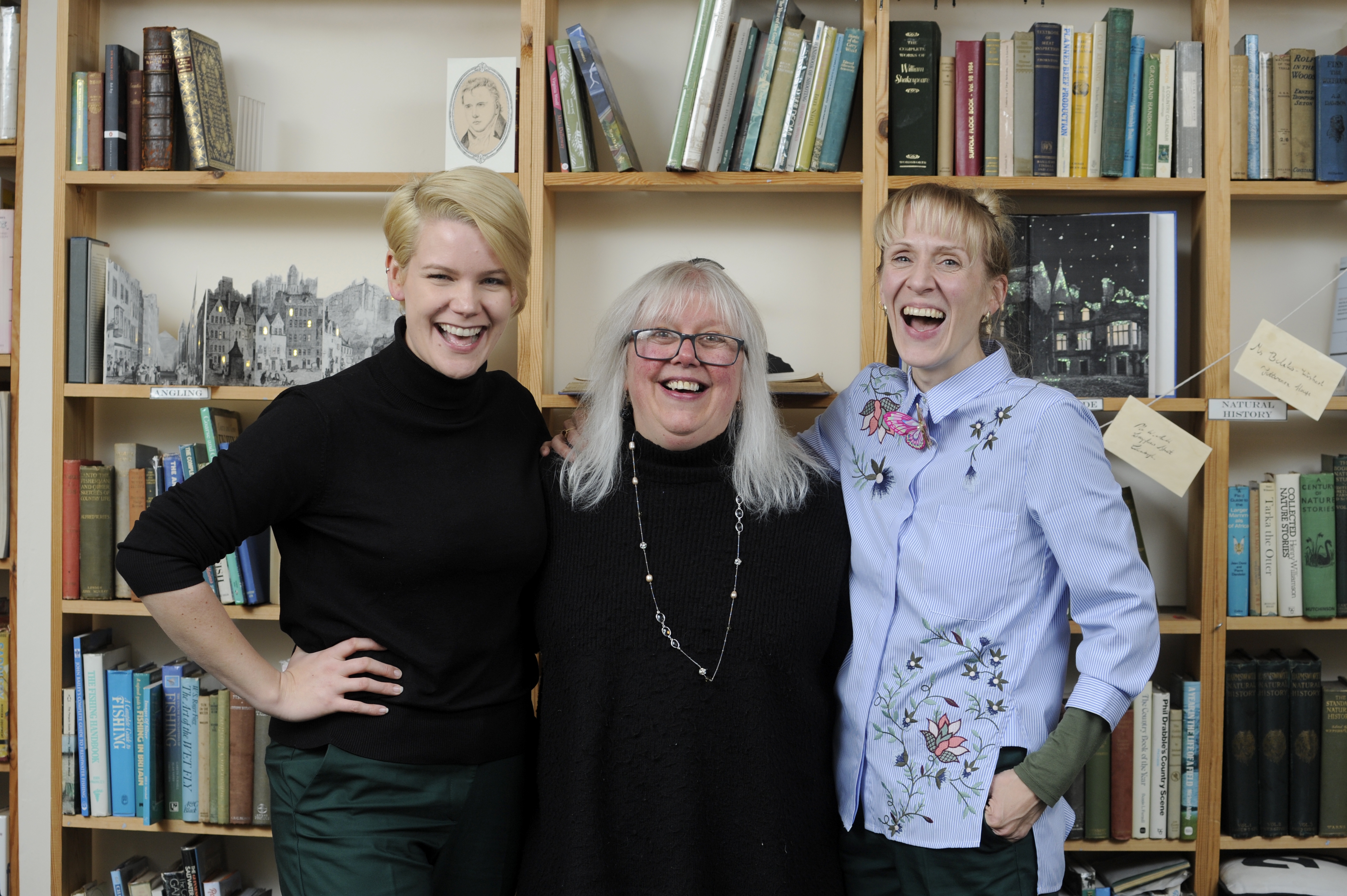 The Old Bank Bookshop owner Joyce Cochrane stands between two people laughing in her bookshop. The bookshelves are full of books.