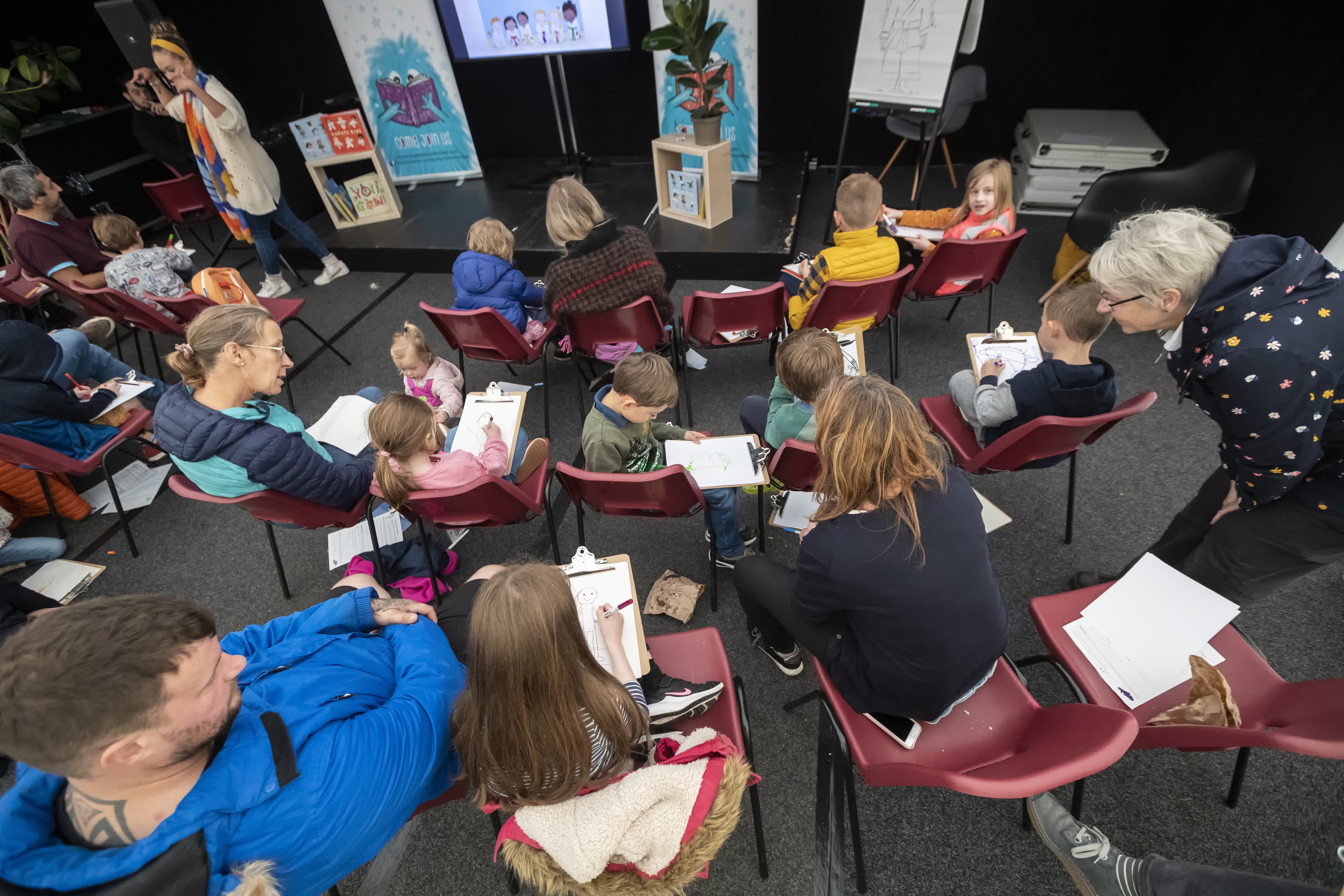 Children and adults sitting in chairs drawing pictures at Holly Stirling's Bigwig event. Holly is talking animatedly in front of the stage.