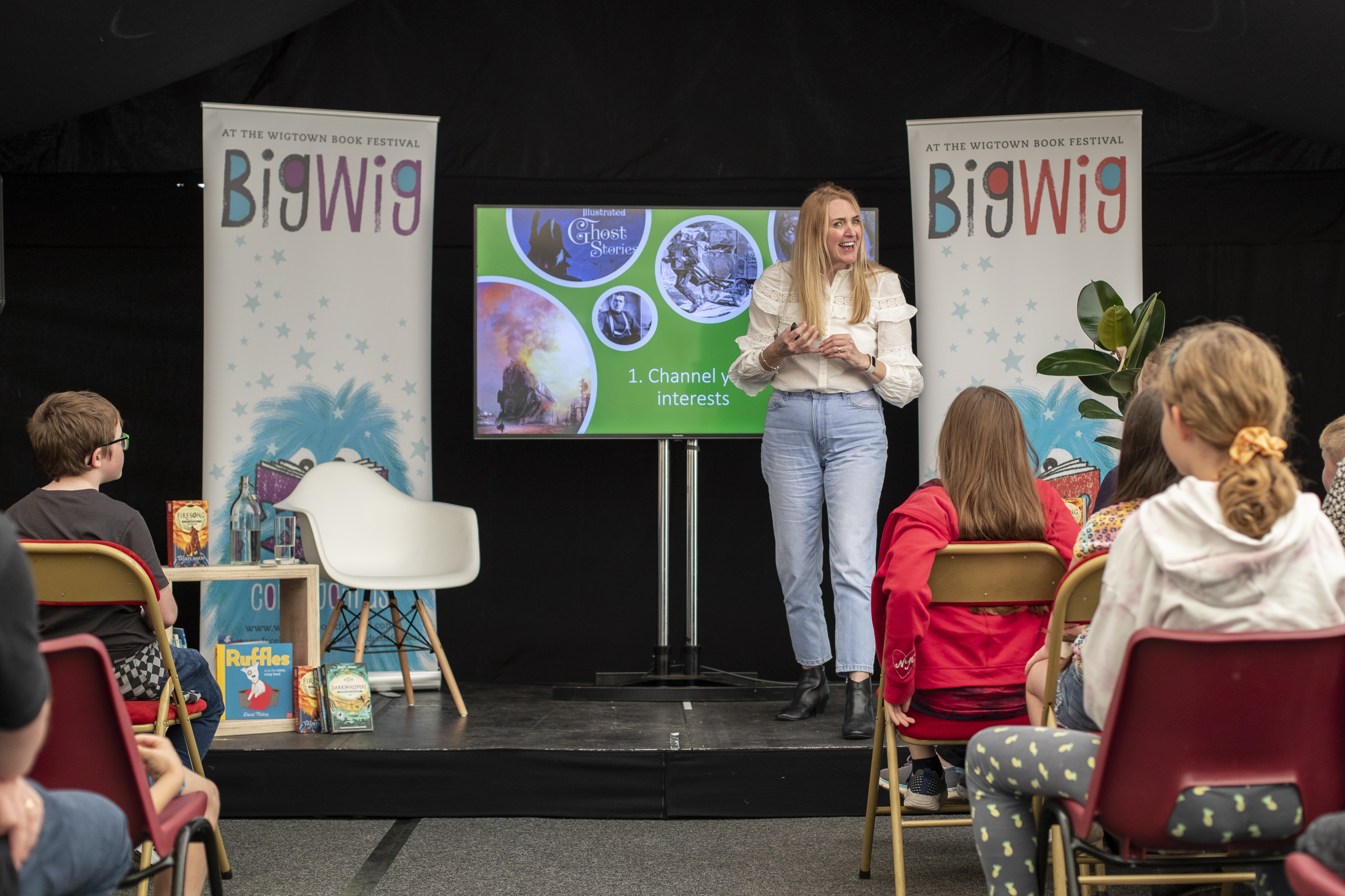 Children's author Vashti Hardy on stage during her event at Wigtown Book Festival. Children are sitting in front of the stage listening. A screen showing images is behind her.