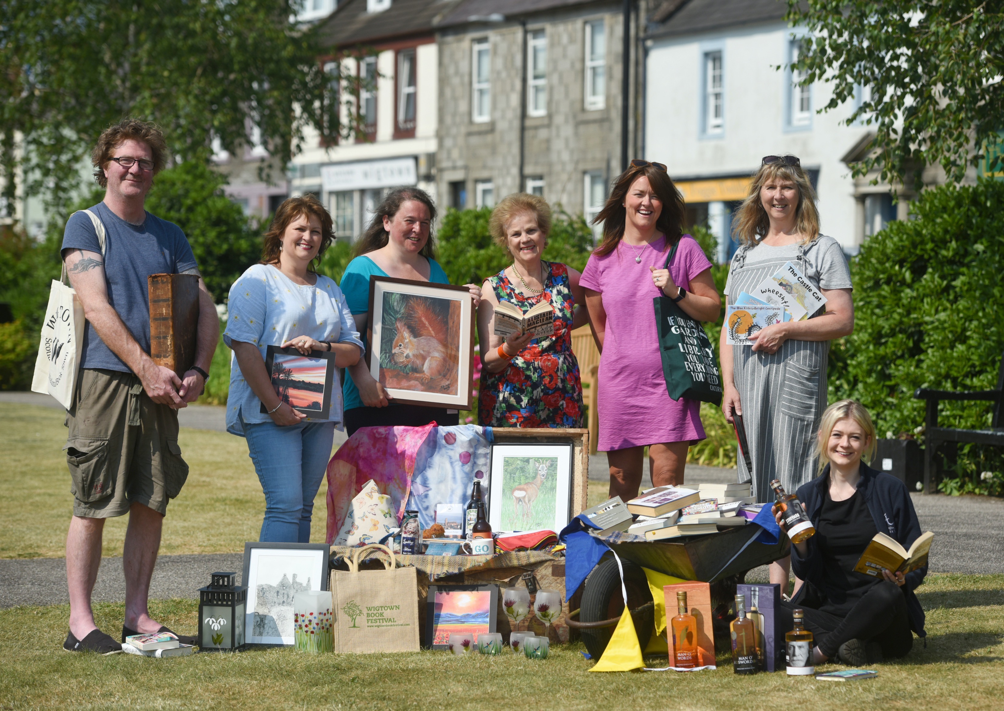 During the Wigtown Book Festival Cathy Agnew, Shaun Bythell and stall holders from The Kist stand together in Wigtown gardens. They are holding books and pictures in front of them are various items.