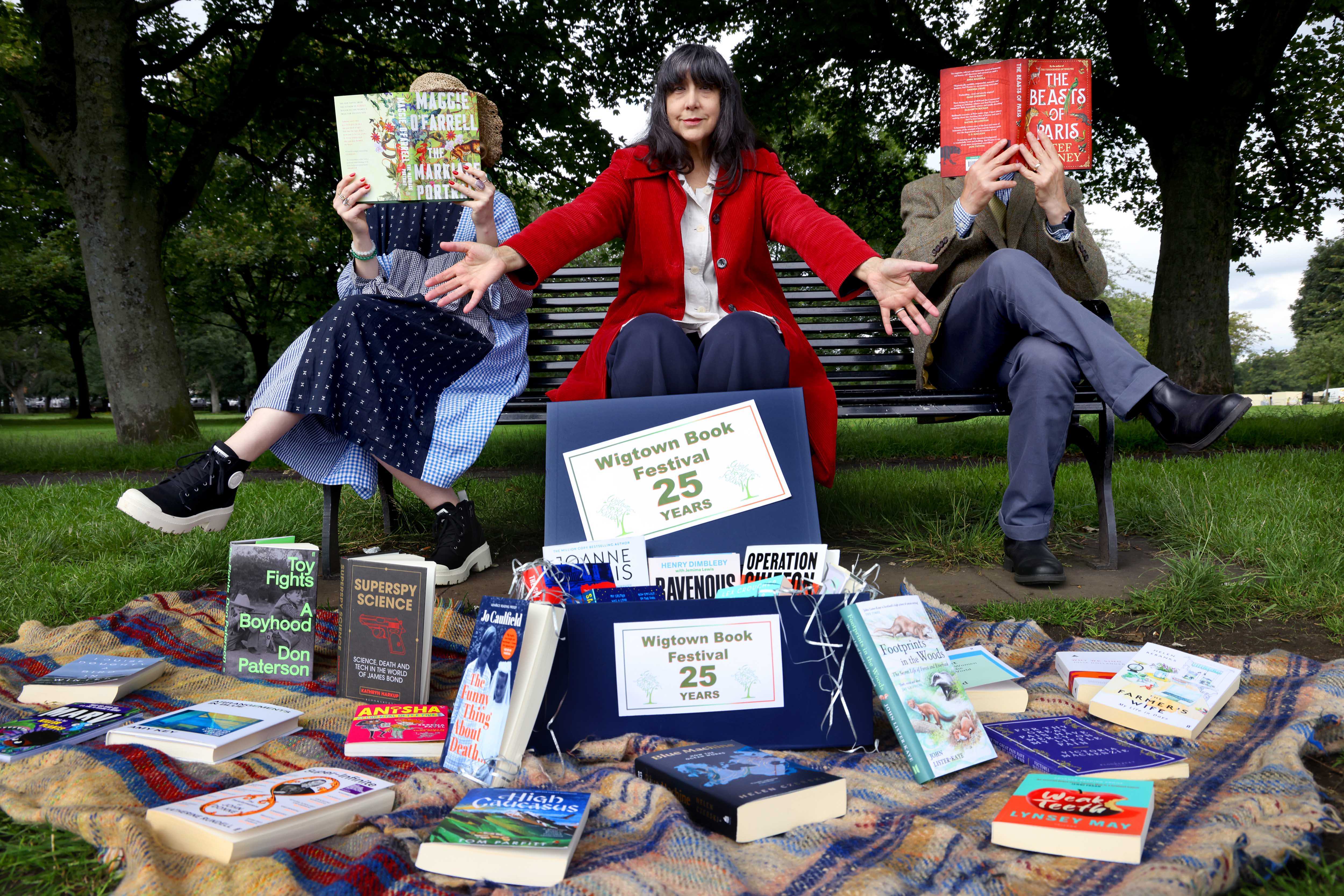 Lee Randall, Guest Programmer at Wigtown Book Festival sitting on a bench in a park. In front of her spread out on a blanket are the 25 year anniversary titles for the book festival. A man and a woman sit either side of her holding up books to obscure their faces.