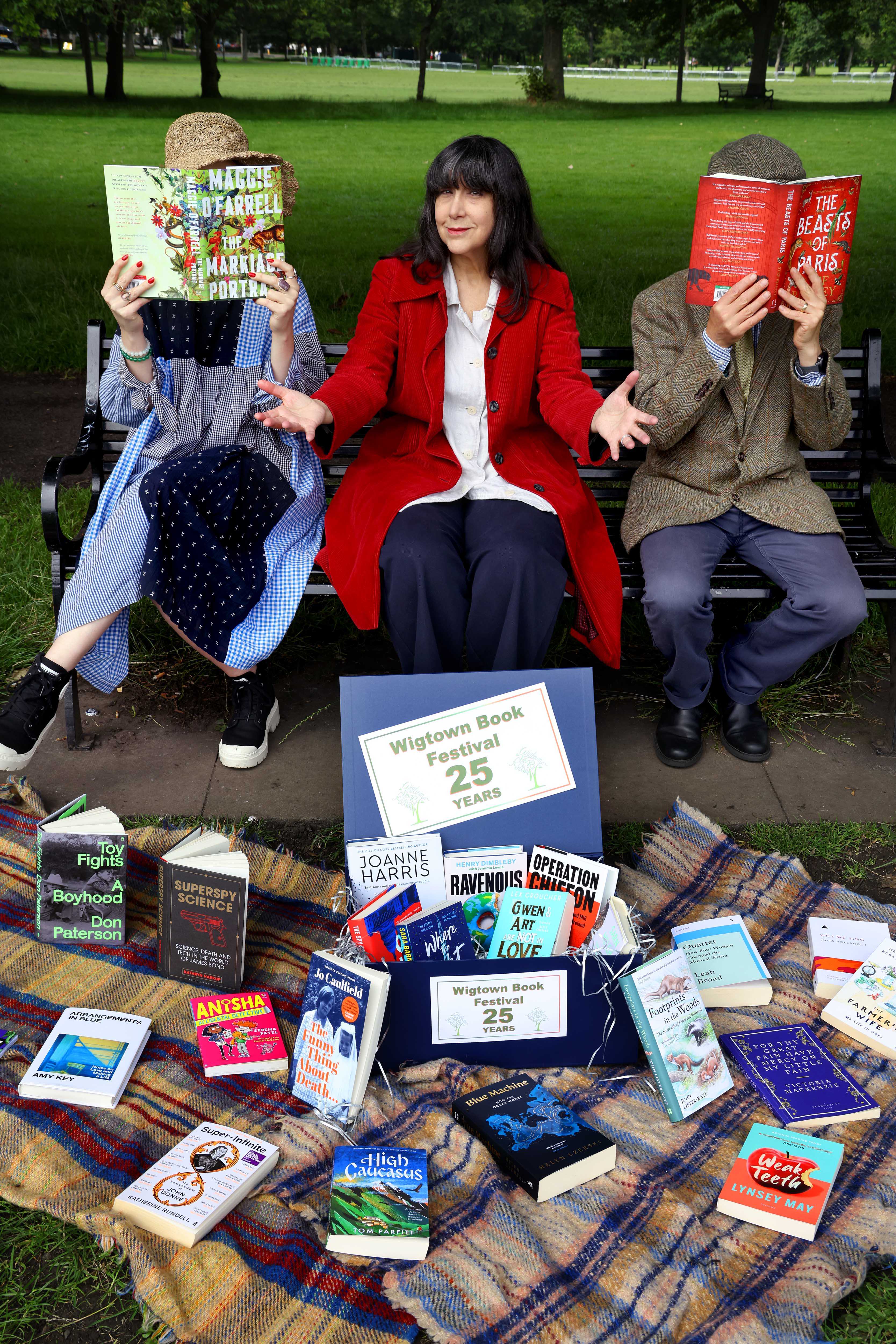 Lee Randall, Guest Programmer for Wigtown Festival Company sits on a bench in a park holding books to promote the twenty fifth anniversary of the Wigtown Book Festival.  On each side of her sits a person also holding an open book, many books are scattered on a blanket in front of them.