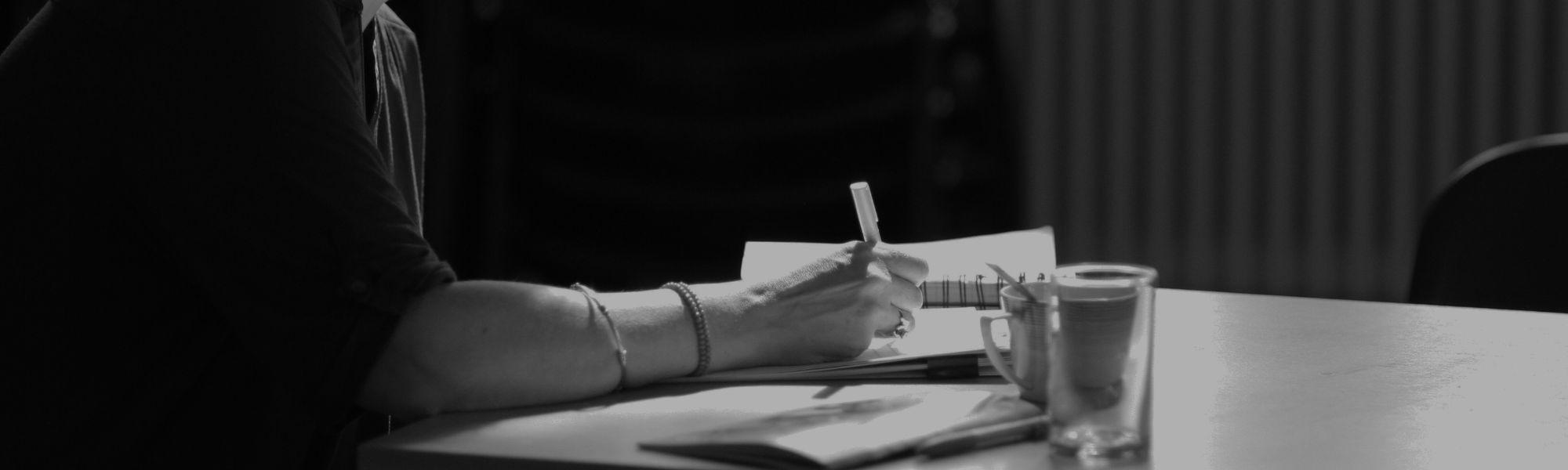 A person sitting at a desk writing in a journal, a small cup and glass beside them.