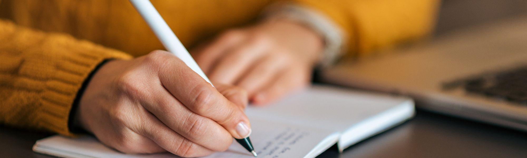 Person wearing  yellow knitted jumper writing in notebook with white pen.