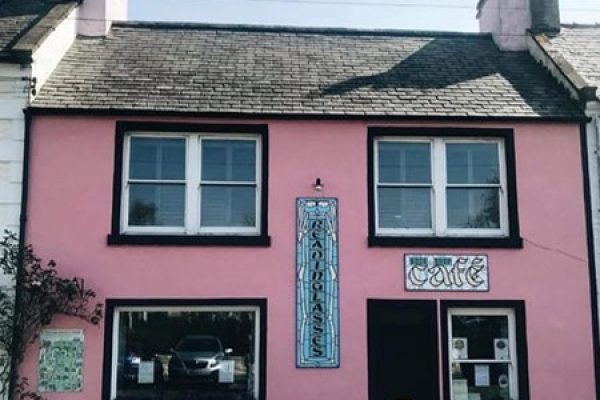 Front view of Cafe Reading Lasses in Wigtown. The house is painted pink with white windows, the cafe sign is vertically placed on the building.