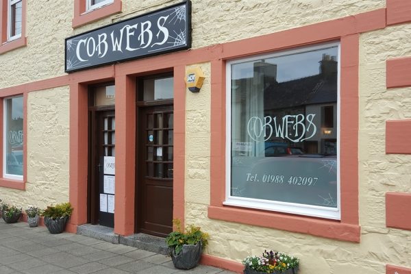 Side view of Cafe Cobwebs in Wigtown. The building is painted yellow with contrasting orange trim around the windows. The cafe sign is above the door in black and white.