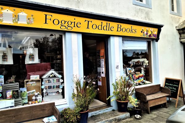 Front view of Foggie Toddle Books in Wigtown.Large windows with the shop name in yellow above. Two benches and pots of flowers outside. Inside the windows are displaying various Wigtown merchandise and books.