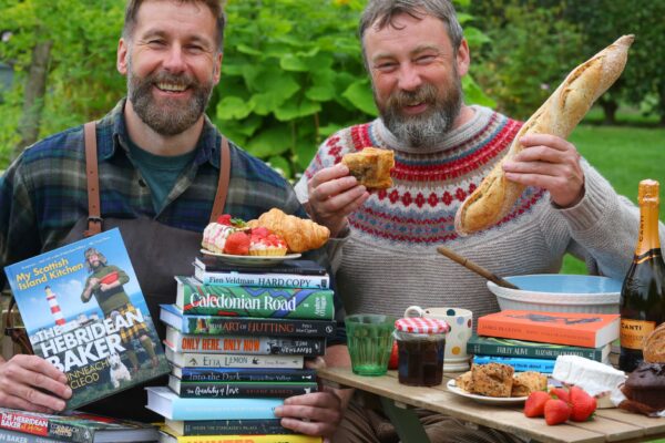 Coinneach MacLeod holding a stack of festival titles on his lap, with a plate of baked goods balanced on top. On the right, Peter MacQueen holding a baguette, both seated at a table containing food, drink and books.
