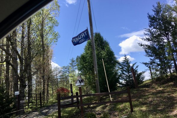 The driveway to a house in North Carolina. A fence, gate and trees line the road. A flag is flying above the brightly coloured mailbox.