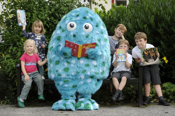 Bigwig, our children's book festival mascot, stands with various children in Wigtown gardens reading a book, The children are smiling and holding books up.