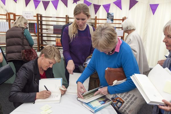 Sports presenter and author Claire Balding is sitting in a chair signing her book in the marquee at a Wigtown Book Festival event. Three people are looking on and talking, bookshelves are in the background with purple bunting hanging up on the wall of the tent.