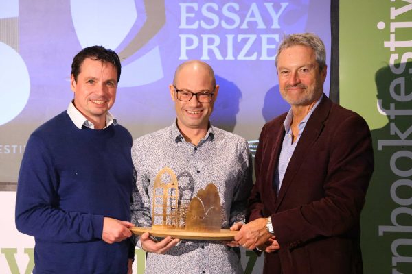 The Anne Brown Prize Winner Rodge Glass stands holding his prize between Adrian Turpin and Gavin Esler, on stage at the Wigtown Book Festival.