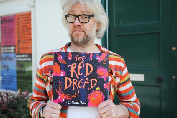 Author Tom Morgan Jones standing in front of  a green door holding his book 'The Red Dread', with a surprised look on his face.