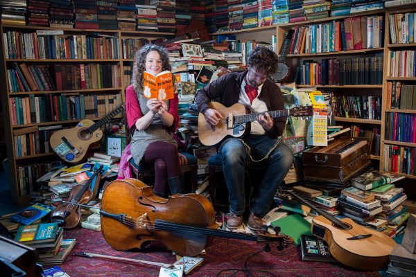 Bookshop band duo Beth and Ben Please sitting in a bookshop surrounded by books. Beth holds a book while Ben plays his guitar.