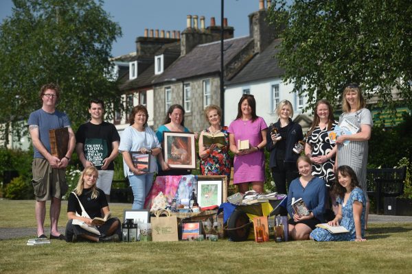 During the Wigtown Book Festival various staff members, bookshop owners and stall holders from The Kist sit and stand together in Wigtown gardens. They are holding books, pictures and bottles of alcohol.