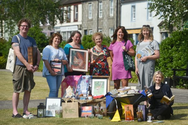 During the Wigtown Book Festival various staff members, bookshop owners and stall holders from The Kist sit and stand together in Wigtown gardens. They are holding books, pictures and bottles of alcohol.