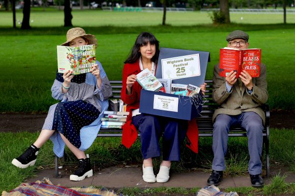 Lee Randall, Guest Programmer for Wigtown Festival Company sits on a bench in a park holding books to promote the twenty fifth anniversary of the Wigtown Book Festival.  On each side of her sits a person also holding an open book.