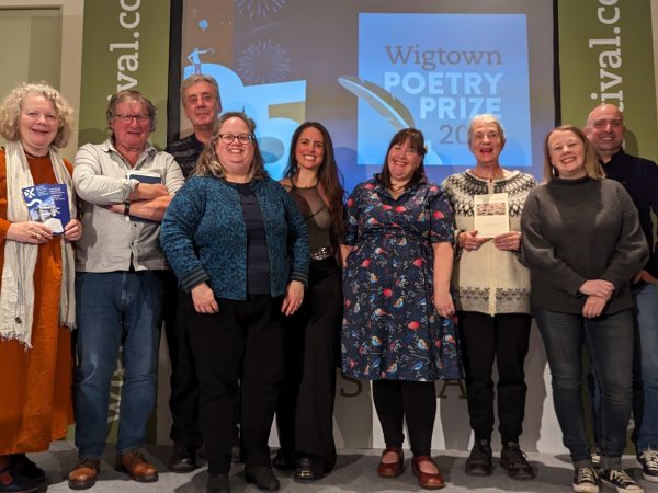 Writers and poetry 2023 winners standing on stage with Poet Hugh McMillan. A screen is behind them showing slideshows.