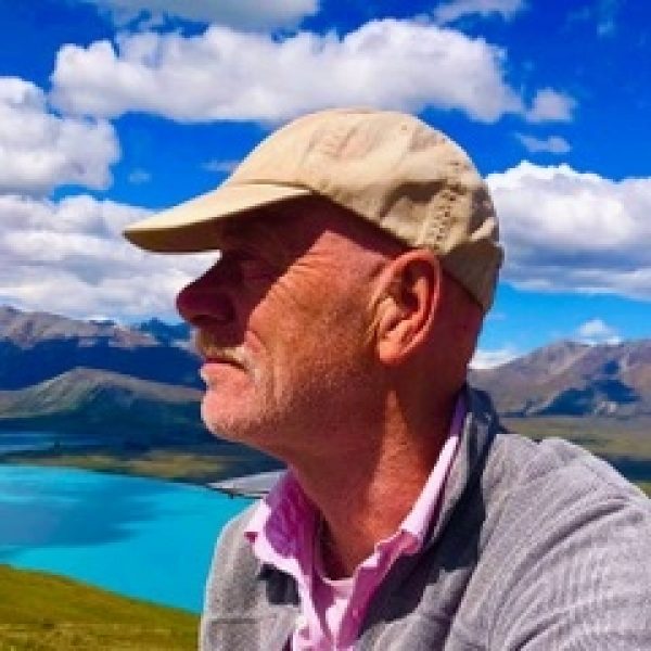 Roger Davis, Vice-Chair at Wigtown Festival Company sitting on a hillside surrounded by mountains and a lake.