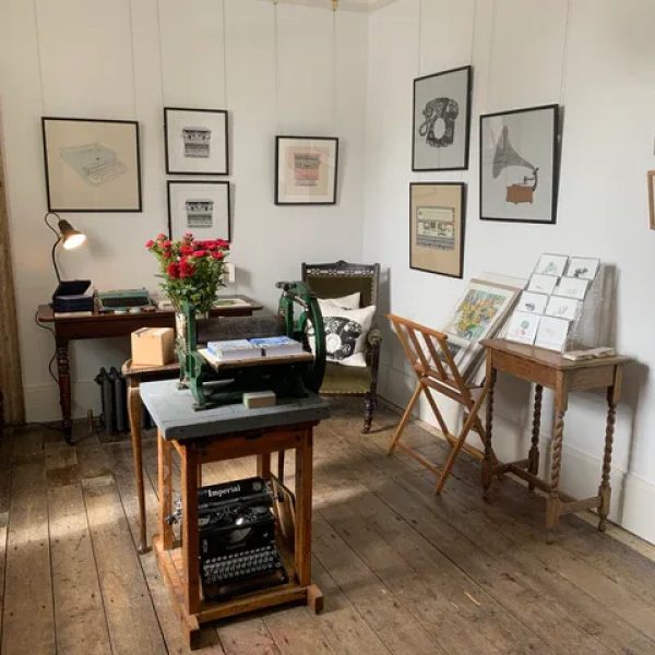 A room inside Craigard Gallery. Painting adorn the walls, three small tables holding various items including cards, a lamp, books and flowers. An old fashioned typewriter sits underneath a small table. The sunshine is streaming through the window.