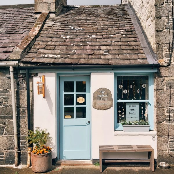 Fountain Stone studio and shop in Bladnoch, Wigtown. A Small terraced cottage painted white with blue window and door. A bench and plant pot sit outside the shop.