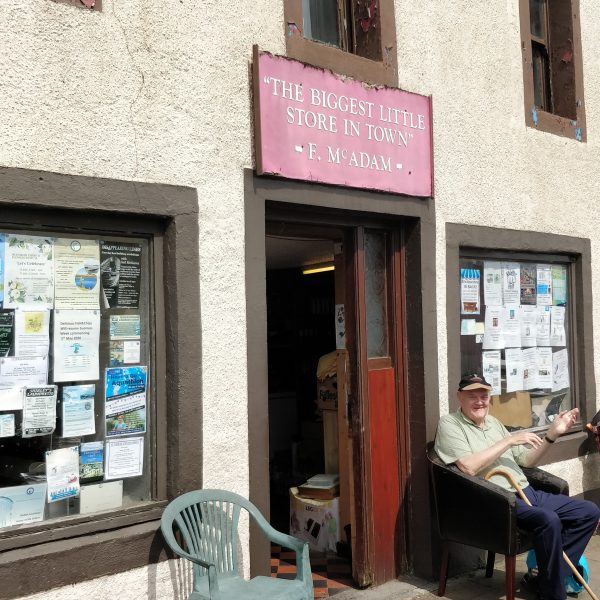 Side view of 'The Biggest little store in town' owned by F McAdam. Cream coloured building with brown windows, covered in various paper notices. Two chairs sit outside, an elderly gentleman sits in one smiling and waving in the sunshine.