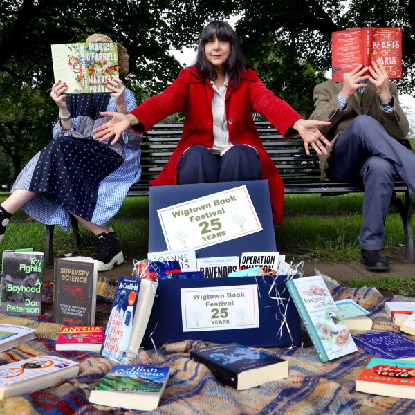 Lee Randall, Guest Programmer at Wigtown Book Festival sitting on a bench in a park. In front of her spread out on a blanket are the 25 year anniversary titles for the book festival. A man and a woman sit either side of her holding up books to obscure their faces.