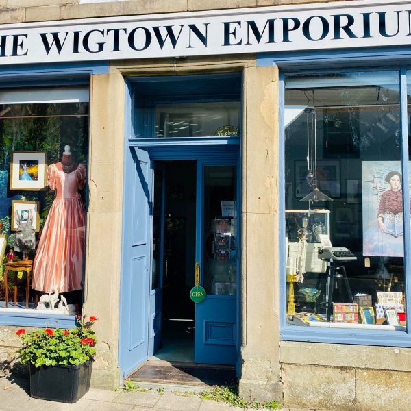 The Wigtown Emporium vintage shop. Large windows display old clothes, books, pictures, typewriters and a lamp. The black and white shop sign sits predominantly above the windows and doors.