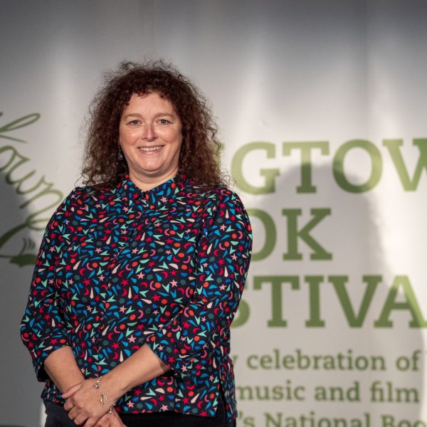 Wigtown Book Festival Poetry Prize winner 2022 Julie Laing. She stands in front of the green and white Wigtown Book Festival logo.