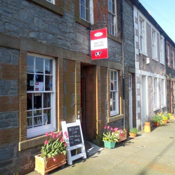 A side view of Beltie Books in Wigtown, Scotland's National Book Town. A terraced stone building with white sash windows. A small A-frame board and plant pots containing tulips sit outside. The red and white shop sign sits above the door.