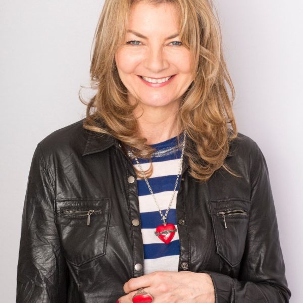 Author jo Caulfield stands smiling wearing a black jacket and blue and white striped t-shirt.