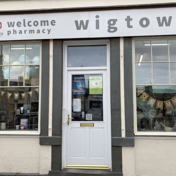 Wigtown pharmacy. A cream coloured building with large windows containing items for sale and bunting. The pharmacy sign sits above the door and windows.