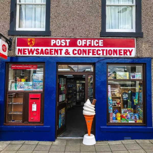 A photograph of Wigtown Post Office before 2020. A pebble dashed building with blue trimmed windows and  post box.